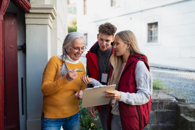 Two young volunteers with a clipboard talking to a senior citizen outside the door to her home.