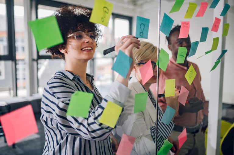 Multiracial team at work writing strategy ideas on sticky notes on the glass wall while having a meeting in the office. Business project planning.