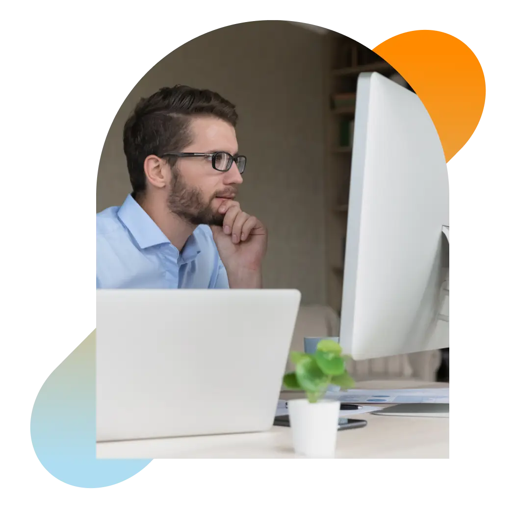 Contemplative man wearing glasses seated in front of computer, staring at large monitor.