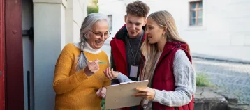 Two young volunteers with a clipboard talking to a senior citizen outside the door to her home.