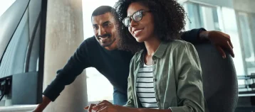 Young businesswoman sitting at her desk working on computer with male colleague standing by